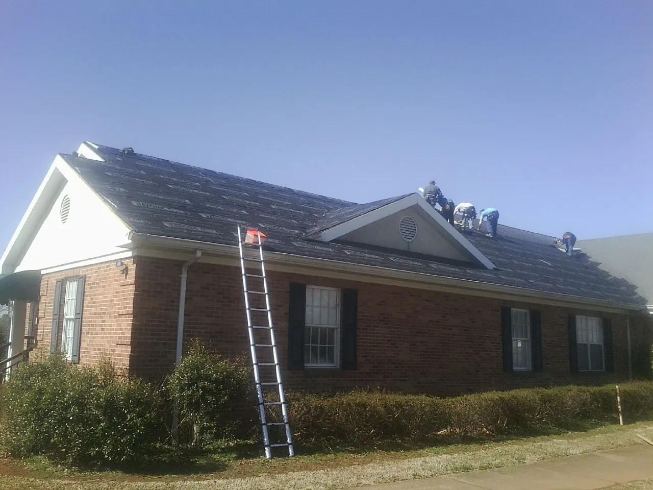 Crew tearing shingles off a brick ranch roof with a ladder set — Emergency Roof Repair work by our crew in New Hudson
