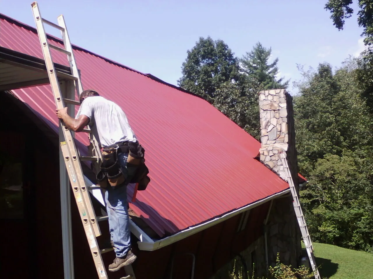 Roofer on a ladder beside a red metal roof and stone chimney — Soffit & Fascia Repair by Roofing Brothers in New Hudson